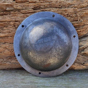 May include: A round, metal shield with a dark grey finish. The shield has a slightly domed shape and is decorated with a series of small, evenly spaced holes around the edge.