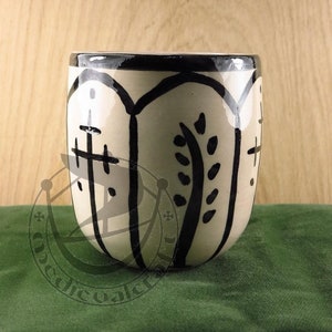 Może przedstawiać: Ceramiczny kubek z kremowym szkliwem i ręcznie malowanym czarnym wzorem. Zewnętrzna strona posiada powtarzające się łukowate panele zawierające stylizowane czarne motywy botaniczne i symbole przypominające krzyże, wykończone czarnym brzegiem.