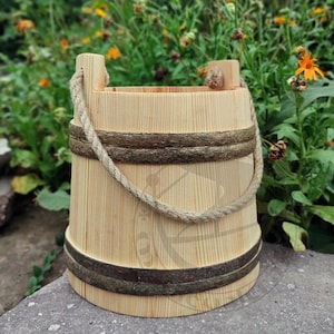 Könnte beinhalten: Ein hellfarbener Holzeimer mit einem Seilgriff. Der Eimer besteht aus vertikalen Holzlatten und hat zwei dunkle Bänder in der Mitte. Der Griff besteht aus einem dicken, gedrehten Seil. Der Eimer steht auf einer Steinoberfläche.