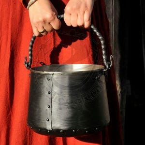 Peut inclure: Un chaudron en métal noir avec une poignée en fer torsadée. Le chaudron a un fond arrondi et est décoré de rivets autour du bord.