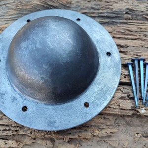 Könnte beinhalten: Ein runder, metallener Schild mit einer gewölbten Mitte und sechs gleichmäßig verteilten Löchern am Rand. Der Schild ist hellgrau und hat eine leicht strukturierte Oberfläche. Fünf Metallnägel liegen auf einer Holzoberfläche neben dem Schild.