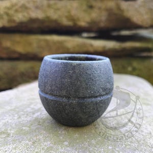 Könnte beinhalten: Ein kleiner, kugelförmiger Becher aus dunkelgrauem Stein mit einer texturierten, matten Oberfläche. Das Gefäß weist eine einzelne horizontale Rille auf, die um die Mitte geschnitzt ist und eine klare Trennung zwischen oberer und unterer Hälfte bildet.