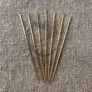 May include: Seven long, slender, gold-coloured needles with small eyelets at the top. The needles are arranged in a fan-like pattern against a textured, neutral-toned fabric background. These sewing needles are designed for various crafting and sewing projects.