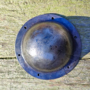 May include: A round, dark gray metal shield with a slightly domed center and a raised rim. The shield has several small holes around the edge.