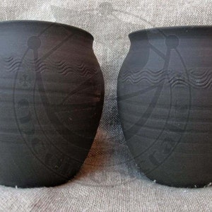 Könnte beinhalten: Zwei schwarze Keramik-Töpfe mit einem wellenförmigen Design um die Mitte. Die Töpfe stehen auf einer strukturierten Stoffoberfläche.