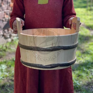 Op de afbeelding: Een lichtgekleurde houten emmer met twee donkere banden en twee houten handvatten. De emmer wordt vastgehouden door een persoon die een rood shirt met lange mouwen draagt. De emmer is gemaakt van verticale houten latten.