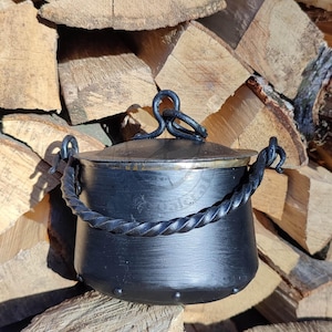 Può includere: Calderone in metallo nero con manico attorcigliato e coperchio. Il calderone ha un bordo in ottone e si trova su una pila di legna da ardere.