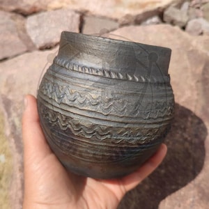 Może przedstawiać: Mały, ciemnobrązowy, zaokrąglony garnek ceramiczny o teksturowanej powierzchni. Zewnętrzna strona posiada poziome pasy z wyrytymi wzorami dekoracyjnymi, w tym rzędem małych pionowych nacięć u góry oraz dwiema wyraźnymi falistymi liniami pośrodku.