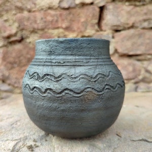 May include: A small, grey ceramic pot with a rounded body and a textured surface. The pot has a wavy, carved design around the middle and horizontal lines near the top. The background is a rough brick wall.