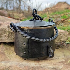 Puede incluir: Caldero de metal negro con tapa y asa de metal retorcido. El caldero tiene una banda de cobre alrededor de la parte superior y está sentado sobre una roca.