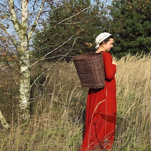 May include: A long-sleeved red dress featuring a dark graphic print near the hem, paired with a white crocheted head covering and a large, deep brown woven wicker basket carried on the back with sturdy straps.