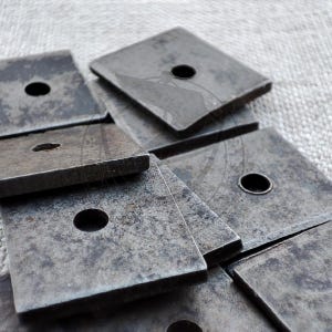 Könnte beinhalten: Ein Stapel quadratischer Metallunterlegscheiben mit einem Loch in der Mitte. Die Unterlegscheiben sind dunkelgrau und haben eine raue Textur.