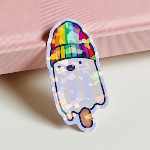 May include: A holographic sticker of a smiling ghost-shaped popsicle wearing a rainbow beanie. The popsicle has a wooden stick and a happy face. The sticker is on a pink surface.