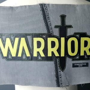 Puede incluir: Un parche de mezclilla gris con la palabra "WARRIOR" en amarillo y negro, junto con un gráfico de espada negra. El parche también incluye el texto "STUBENSPHINX DENIM".