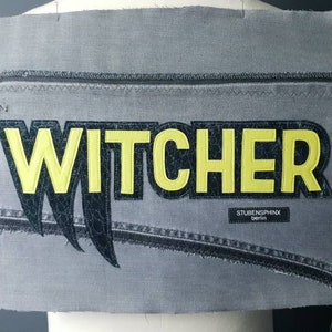 Könnte beinhalten: Ein graues Denim-Patch mit dem Wort "WITCHER" in gelben Buchstaben und schwarzen Krallen darunter. Der Patch enthält auch den Text "STUDIOSPHINX berlin".