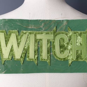 Könnte beinhalten: Ein grünes Stoffpatch mit dem Wort "WITCH" in hellgrünen, stilisierten Buchstaben. Das Patch ist rechteckig mit einem gezackten Randdesign. Auf der rechten Seite befindet sich ein kleines schwarzes Etikett mit weißem Text.