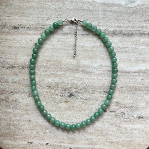 May include: A necklace composed of smooth, round, light green beads arranged in a circular pattern. The necklace features a silver-toned metal lobster claw clasp and an adjustable chain extender at the top, providing a secure closure mechanism.