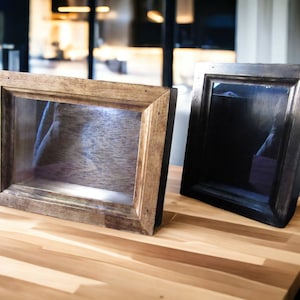 Könnte beinhalten: Zwei Holz-Schattenboxen mit Glasfronten. Die Box auf der linken Seite ist hellbraun mit einer natürlichen Holzoptik. Die Box auf der rechten Seite ist dunkelbraun mit einer schwarzen Lackierung.