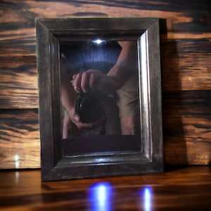 Könnte beinhalten: Ein schwarzer Spiegelrahmen mit der Reflexion einer Person darin. Die Person hält eine Kamera in der Hand. Der Spiegel steht auf einer Holzoberfläche.