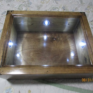May include: A wooden display case with a glass top and a wood frame. The case has a light brown finish and is illuminated by five small white lights.