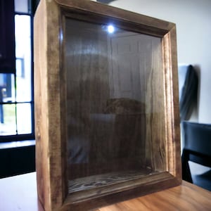 Puede incluir: Una caja de sombra de madera con un frente de vidrio transparente y una luz en el interior. La caja está sentada sobre una mesa de madera.