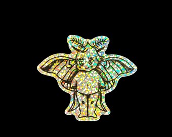 May include: A holographic sticker featuring a cartoonish winged creature with a bow, horns, and a smiling face. The sticker has a glittery, iridescent finish, with black outlines and details. The background is solid black.