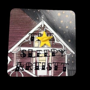 May include: A square sticker with a detailed illustration of a house. The house has a burgundy roof and white trim. The text "THE SLEEPY ARTIST" is displayed on the house. A yellow star is affixed to the top. The background is a night sky.
