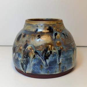 May include: A round ceramic vase with a wide base and a narrow neck. The glaze features a blend of deep blue, tan, and dark brown tones with a dripped, mottled texture that transitions from a lighter top to a darker, saturated blue base.