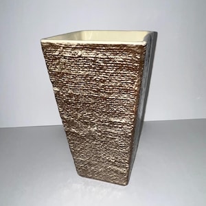 May include: A square-shaped vase with a tapered base and a wide opening. The exterior features a textured, dark brown finish with horizontal ridges, while the interior and the top rim are finished in a smooth, solid off-white color.