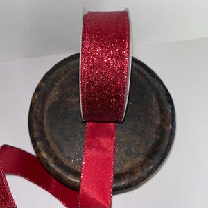 Puede incluir: Un rollo de cinta roja con purpurina y borde de sat&eacute;n. La cinta se muestra sobre una superficie oscura y redonda. La purpurina capta la luz, creando un efecto brillante. La cinta est&aacute; desenrollada, mostrando el borde de sat&eacute;n.