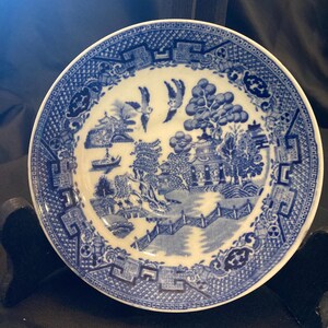 Op de afbeelding: Een klein, rond, blauw en wit decoratief bord met een klassiek Willow-patroon. Het bord toont een gedetailleerde landschapsscène met pagodes, bomen, een brug en vogels. De rand heeft een geometrische rand.