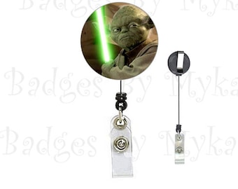 May include: A green and brown Yoda character from Star Wars holding a green lightsaber. The image is on a round badge holder with a black retractable cord and a silver clip.
