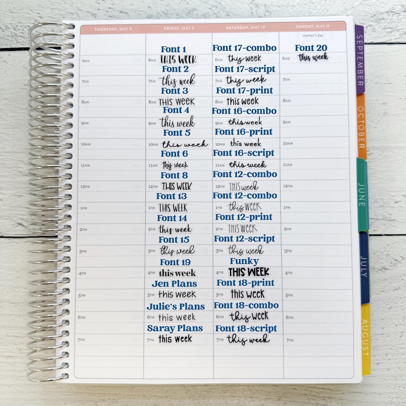 May include: A spiral-bound planner with a white interior, featuring a weekly schedule layout. The pages are filled with handwritten text in various fonts, including the words "THIS WEEK" and the names of fonts. Colorful tabs mark the months.