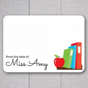 May include: A white rectangular desk nameplate with rounded corners. The text "From the desk of: Miss Amy" is printed in black. A red apple and a stack of books in green, blue, and red are on the right.