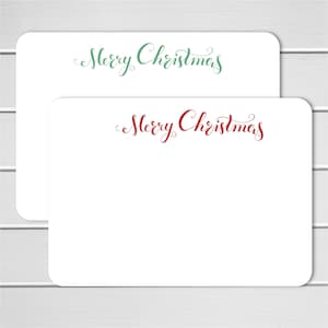 May include: Two white rectangular cards with rounded corners. The top card has "Merry Christmas" in green script, and the bottom card has the same text in red script. The cards are on a white wooden surface.