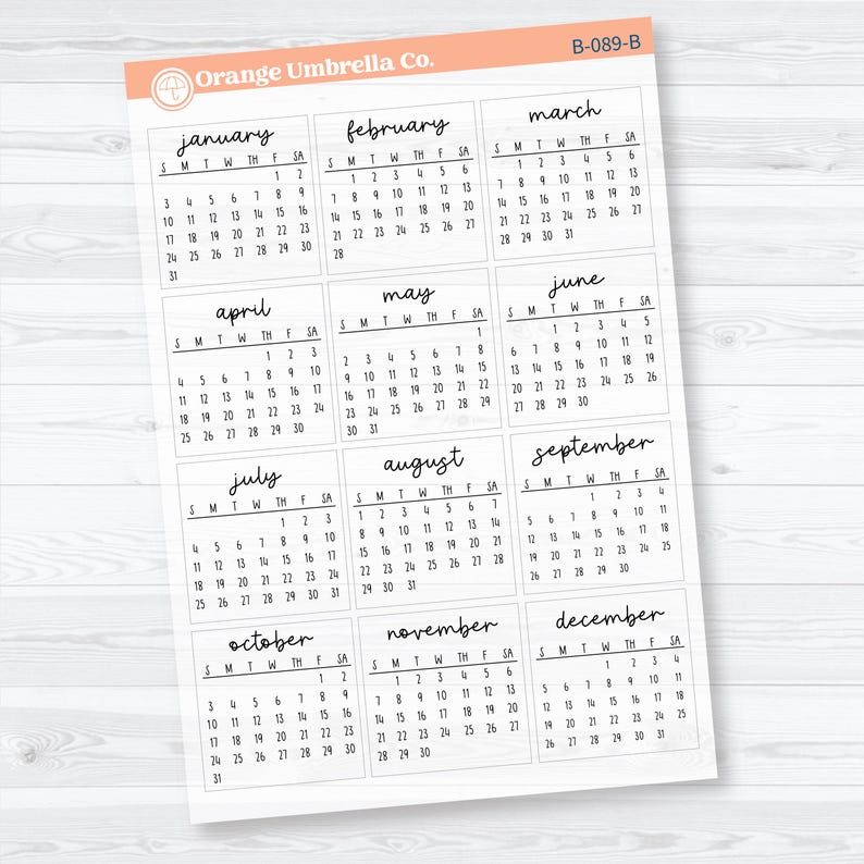 Op de afbeelding: Een witte papieren kalender met maandelijkse kalenderindelingen in handschrift. Elke maand is benoemd. Het logo van Orange Umbrella Co. staat linksboven. Afmetingen in centimeters.
