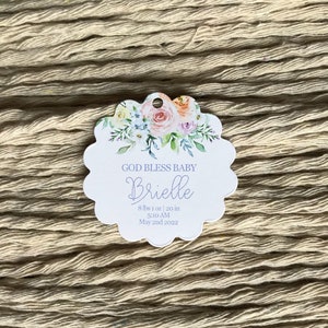 Op de afbeelding: Een geschulpte tag met bloemaccenten en de tekst "GOD BLESS BABY Brielle". De tag bevat de geboortegegevens van de baby: 3,65 kg, 50,8 cm, 5:10 uur en 2 mei 2022. De achtergrond is een getextureerd, beige oppervlak.