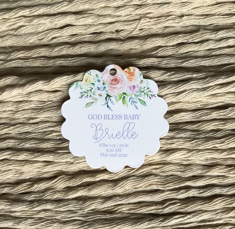 Op de afbeelding: Een geschulpte tag met bloemaccenten en de tekst "GOD BLESS BABY Brielle". De tag bevat de geboortegegevens van de baby: 3,65 kg, 50,8 cm, 5:10 uur en 2 mei 2022. De achtergrond is een getextureerd, beige oppervlak.