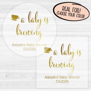 May include: A clear acrylic circle with gold foil text that reads "a baby is brewing" and "Aaliyah's Baby Shower 5.9.2026". The text is in a cursive font. There is a small image of a teacup on the left side of the circle. The circle is on a white wooden background.