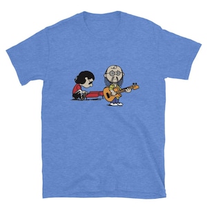 May include: Blue short-sleeve t-shirt featuring a cartoon illustration of two figures. One figure is seated and playing a red instrument, while the other stands and plays a guitar. The figures are in a cartoon style.