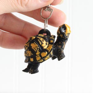 May include: A black and gold turtle keychain with a silver keyring. The turtle figurine has a detailed shell pattern and a yellow head. The keychain is held against a white background.