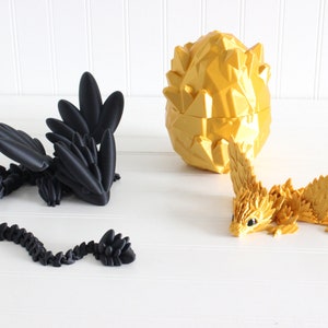 Peut inclure: Deux figurines de dragons, l'une noire et l'autre dorée, avec un œuf de dragon doré en arrière-plan. Les dragons sont faits d'un matériau texturé et ont des ailes et des queues détaillées.
