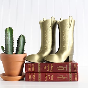 May include: Two gold cowboy boot bookends on top of two red hardback books. The books are titled "The Saddle and Border Trumpet Ride" and "Ernest Haycox". A potted cactus is in the foreground.