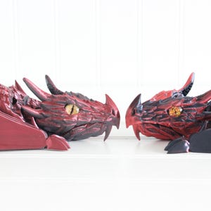 Peut inclure: Deux figurines de dragons rouges et noirs se faisant face. Le dragon de gauche a un corps rouge et des ailes noires, tandis que le dragon de droite a un corps noir et des ailes rouges. Les deux dragons ont des yeux dorés.