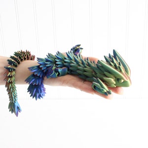 Peut inclure: Une figurine de dragon iridescente verte et bleue avec un design épineux et écailleux. Le dragon est en position recroquevillée, la tête reposant sur ses pattes avant.