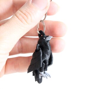 May include: A black raven keyring with a silver ring. The raven has a detailed design with grey accents on the beak and feet. The keyring is held against a white background.