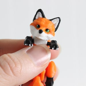 May include: A small, orange and white fox figurine with black accents on the ears and paws. The fox has green eyes and is held in a person's hand. The figurine is a miniature representation of a fox.