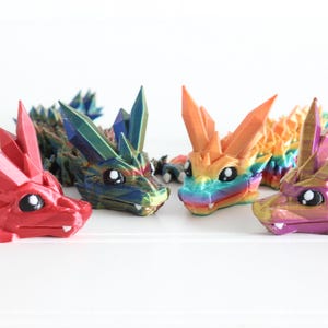 Peut inclure: Plusieurs figurines de dragons colorés et géométriques. Les dragons présentent des structures cristallines pointues dans des tons de rouge, bleu, orange et violet. Chaque dragon a de grands yeux ronds et un corps détaillé.