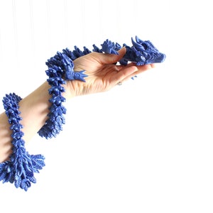 Peut inclure: Un bracelet en plastique bleu en forme de dragon avec une tête et des écailles détaillées. Le bracelet est flexible et peut être porté au poignet.
