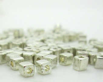 May include: A close-up of a pile of silver square beads. The beads are small and have a hole in the center.